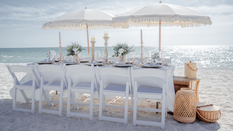 Sunlit seaside dining setup on white sand — long table with white folding chairs, fringed umbrellas, floral centerpieces, candles, glassware and wicker accents by the sparkling ocean