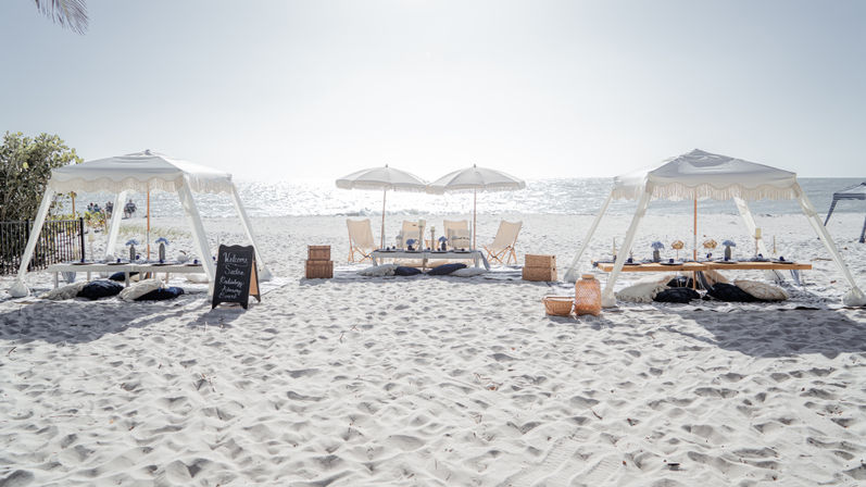 White-sand beach picnic setup with fringed cabanas, umbrellas, low tables, cushions and wicker baskets facing a sparkling ocean view
