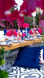 Vibrant outdoor low-table setup with blue velvet floor cushions, wooden table dressed in blue-and-white china, rose-gold wine glasses, bright pink floral centerpieces and pink candlesticks on a patterned rug
