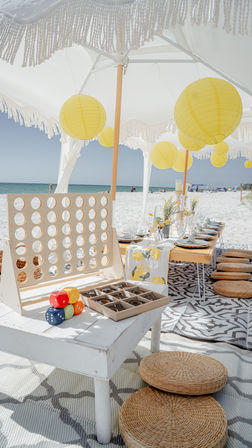 Boho coastal picnic on white-sand beach with fringed umbrella and yellow paper lanterns, low wooden table set for a seaside brunch, woven floor cushions, tabletop games and ocean horizon