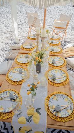 Beach picnic table on white sand with lemon-themed plates, yellow woven chargers, candles, glassware and cozy cushions for sunny coastal outdoor dining
