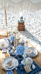 Boho-chic beach picnic tablescape on white sand under a fringed umbrella with blue hydrangeas, wooden candleholders, seashell decor, gold cutlery and blue glassware.
