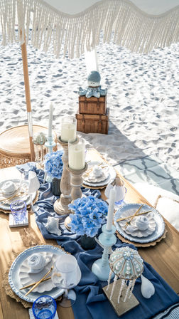 Boho-chic beach picnic tablescape on white sand under a fringed umbrella with blue hydrangeas, wooden candleholders, seashell decor, gold cutlery and blue glassware.