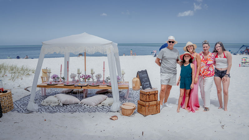 Multigenerational family posing beside a boho beach picnic on white sand — fringe canopy, low table set with candles and florals, wicker baskets and a chalkboard birthday sign — calm blue ocean and horizon in the background.