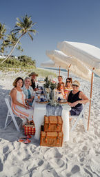 Six friends enjoying a styled beach picnic on a white‑sand shore under fringed umbrellas, palm trees and ocean in the background, elegant table setting and wicker baskets