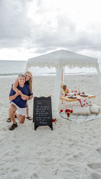 Smiling couple embracing at a romantic beach picnic under a white fringed canopy, low wooden table set with plates and red roses, chalkboard sign on sandy shore with ocean and cloudy sky.
