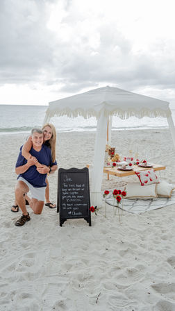 Smiling couple embracing at a romantic beach picnic under a white fringed canopy, low wooden table set with plates and red roses, chalkboard sign on sandy shore with ocean and cloudy sky.