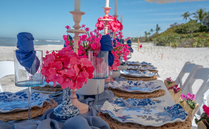 Vibrant beachside tablescape on white sand with blue-and-white floral plates, woven chargers, navy napkins in glassware, bright pink floral centerpieces and candlesticks, with ocean waves and palm trees in the background