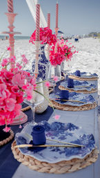 Beachside table on white sand with blue-and-white floral plates, woven placemats, gold cutlery, navy napkins, wine glasses, vibrant pink flower centerpieces and tall twisted pink candles, ocean in the background
