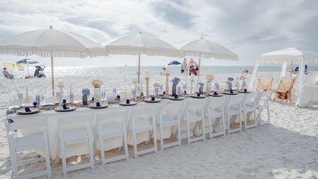 Long white banquet table set for a beachside dinner on powdery white sand, navy place settings and candles under fringe umbrellas with the ocean horizon and a distant sailboat