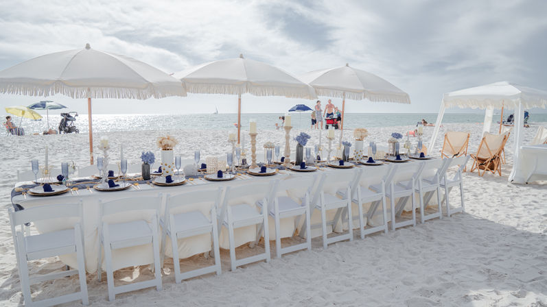 Long white banquet table set for a beachside dinner on powdery white sand, navy place settings and candles under fringe umbrellas with the ocean horizon and a distant sailboat