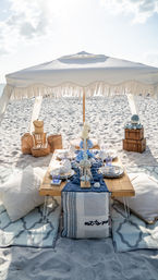 Boho seaside picnic on a sandy beach under a white fringed umbrella — low wooden table with blue-and-white linens, plates, glassware, candles, wicker baskets, stacked trunks and floor cushions.