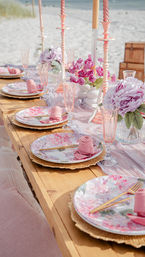 Seaside tablescape on a wooden table with pastel pink floral plates, rolled rose napkins, gold cutlery, pink glassware, tall candles and peony bouquets, sandy beach and ocean in the background.