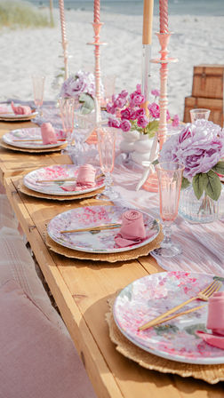 Seaside tablescape on a wooden table with pastel pink floral plates, rolled rose napkins, gold cutlery, pink glassware, tall candles and peony bouquets, sandy beach and ocean in the background.