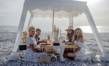 Smiling woman and three children enjoying a boho canopy picnic on a sandy oceanfront at sunset, seated around a low wooden table set with candles, glassware and plates