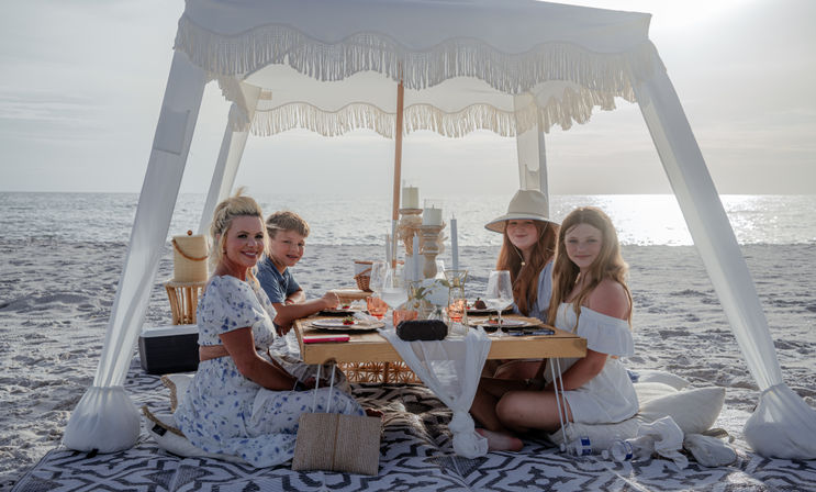 Smiling woman and three children enjoying a boho canopy picnic on a sandy oceanfront at sunset, seated around a low wooden table set with candles, glassware and plates