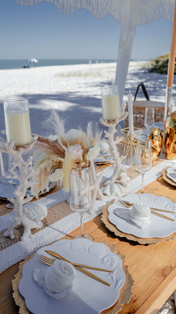 Sunny beachfront table setting on white sand — boho coastal décor with scalloped shell plates on gold chargers, gold cutlery, rolled linen napkins, coral candleholders, pampas grass and champagne flutes.