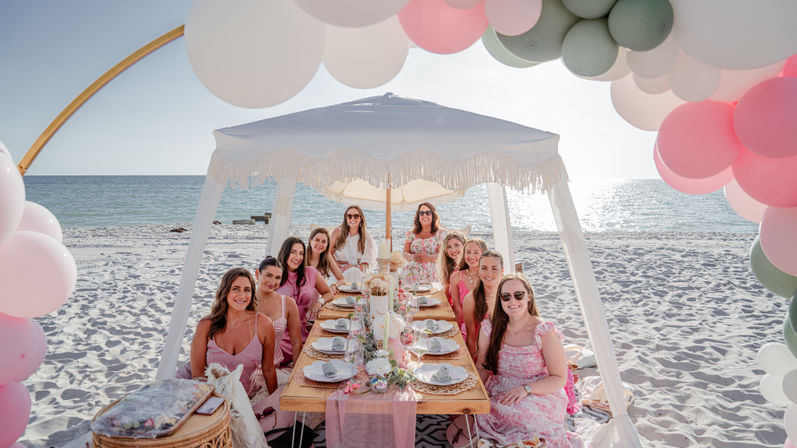 Group of women enjoying a boho beach picnic under a fringed white canopy on a sunlit sandy shore, seated at a low wooden table with pink-and-green balloon arch, floral tablescape and place settings by the ocean