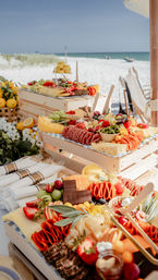 Sun-drenched beachfront charcuterie spread on wooden crates with cheeses, cured meats, fresh fruit, olives and chocolate overlooking white sand and turquoise ocean.