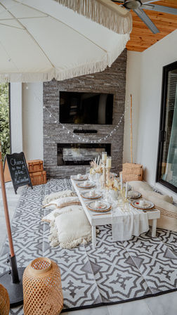 Boho-chic covered patio dining setup with low white table, floor cushions on a geometric rug, stone fireplace, fringe umbrella, string lights and rattan lanterns