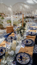 Sunlit boho beach picnic on white sand under a fringed umbrella — low wooden table with blue patterned plates, gold flatware, navy napkins and floral centerpieces.