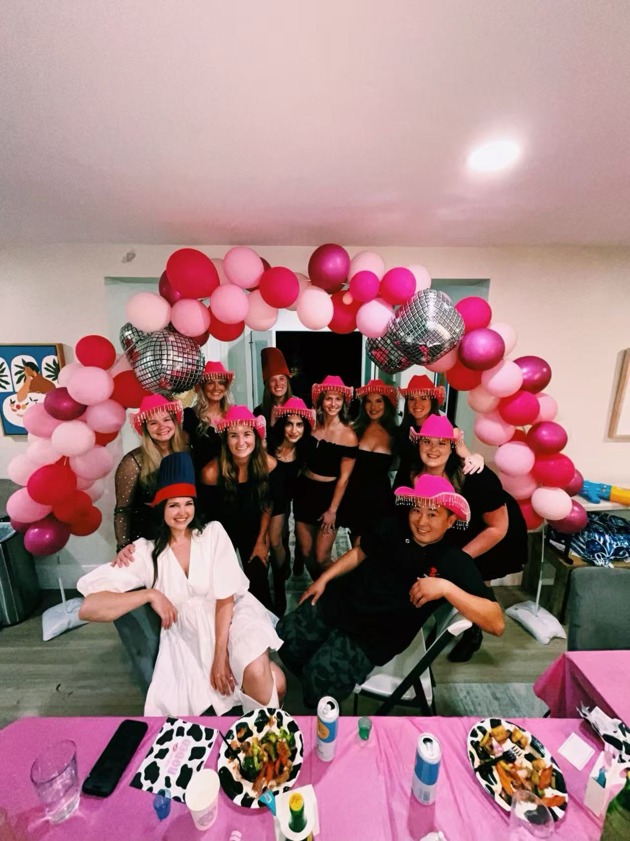 Indoor celebration with a group of friends posing under a pink and magenta balloon arch with disco-ball accents; many wear bright pink fringe party hats, a woman in a white dress sits in front, and a table with plates, drinks and a pink tablecloth fills the foreground.