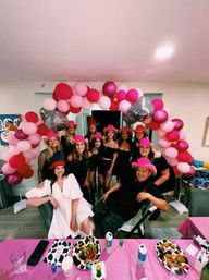 Group of friends at an indoor house party posing under a pink and red balloon arch with mirrored disco balls, wearing pink fringe hats and smiling behind a table of plates and drinks.