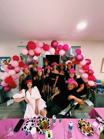 Indoor celebration with a group of friends posing under a pink and magenta balloon arch with disco-ball accents; many wear bright pink fringe party hats, a woman in a white dress sits in front, and a table with plates, drinks and a pink tablecloth fills the foreground.