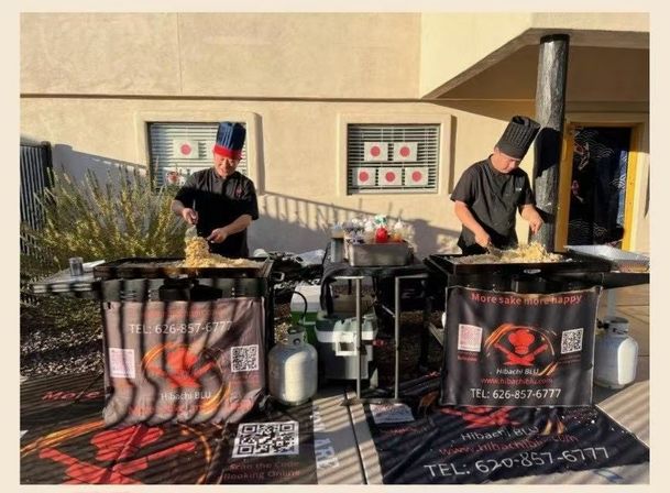 Two chefs sizzling fried rice on outdoor hibachi flat-top grills at a mobile pop-up station, flanked by propane tanks, condiments, and promotional banners.