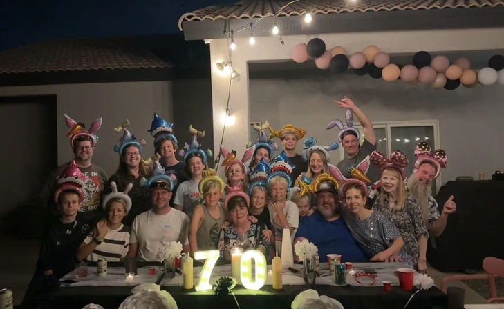 Multi-generation family and friends posing at a backyard patio birthday party at night, wearing colorful animal and unicorn hats under string lights and a balloon garland, with illuminated "70" marquee on a decorated table.