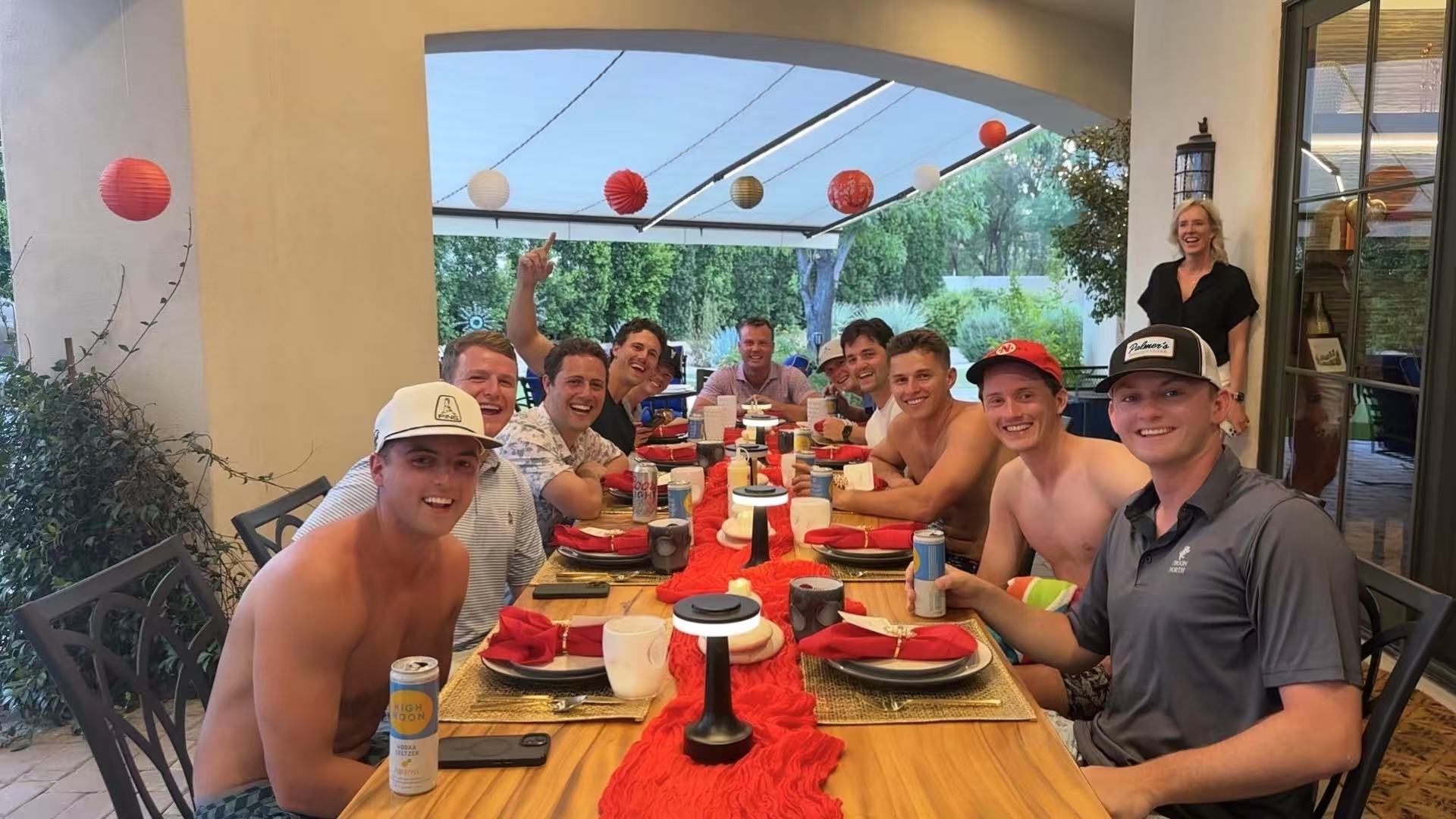 Group of young men and a woman laughing around a long covered backyard patio table set with red table runners, place settings, drinks and hanging paper lanterns for a summer dinner