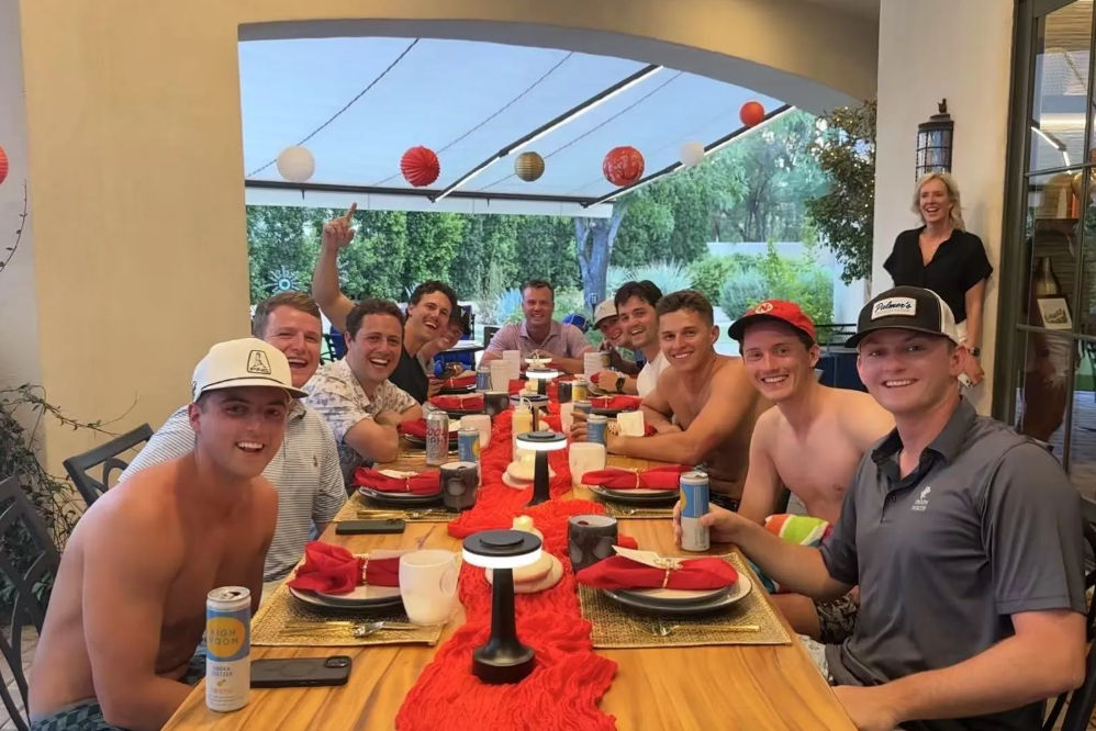 Group of friends gathered around a long wooden dining table on a covered backyard patio, smiling and holding drinks with red table runner, place settings and hanging paper lanterns — outdoor summer dinner party.