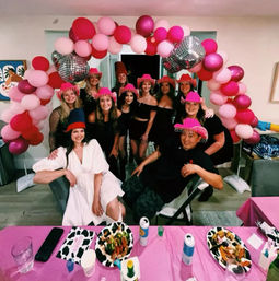 Cheerful group of friends posing under a pink and magenta balloon arch in a living room, wearing playful fringe hats with plates of party food and cans of drinks on a pink-clothed table — indoor celebration.