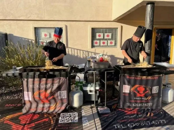 Two cooks in tall hats stir-frying noodles on large outdoor hibachi griddles at a sunny pop-up food stall with propane tanks and promotional banners.