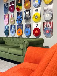 Colorful vinyl record wall display above a green velvet sofa and bright orange armchair in a modern music lounge.