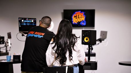 Behind-the-scenes shot of a DJ mixing session in a modern recording/control room — two people seen from behind at a laptop running DJ software, ring light, wall screen with a colorful logo, and a yellow-cone studio monitor speaker.