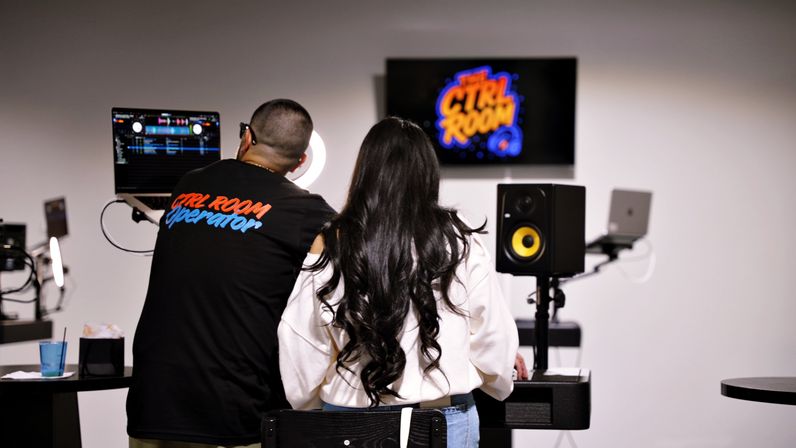 Behind-the-scenes shot of a DJ mixing session in a modern recording/control room — two people seen from behind at a laptop running DJ software, ring light, wall screen with a colorful logo, and a yellow-cone studio monitor speaker.