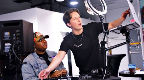 Standing DJ adjusts laptop and ring light while coaching a seated person at a mixing controller in a bright indoor studio livestream setup.