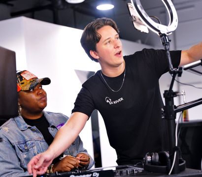 Standing DJ adjusts laptop and ring light while coaching a seated person at a mixing controller in a bright indoor studio livestream setup.