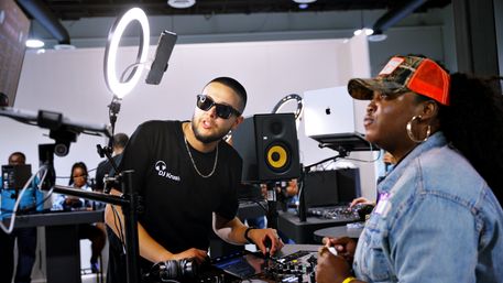 Two DJs at an indoor music booth — a man in sunglasses adjusts a digital mixer under a ring light and studio monitor while a woman in a red trucker cap and denim jacket watches nearby