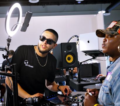 Two DJs at an indoor music booth — a man in sunglasses adjusts a digital mixer under a ring light and studio monitor while a woman in a red trucker cap and denim jacket watches nearby