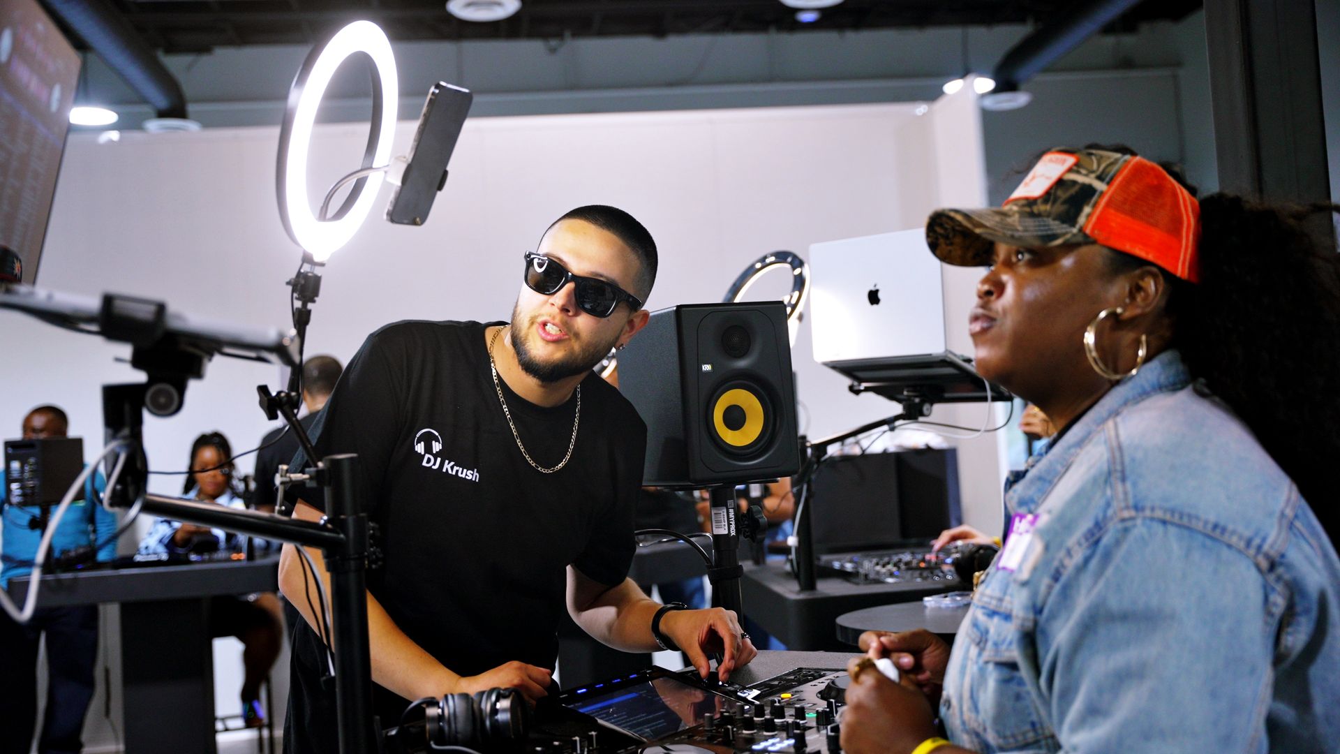 Two DJs at an indoor music booth — a man in sunglasses adjusts a digital mixer under a ring light and studio monitor while a woman in a red trucker cap and denim jacket watches nearby