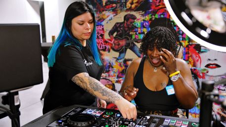Two women at a DJ controller in a bright, colorful music studio — one demonstrates the controls while the other laughs under a ring light