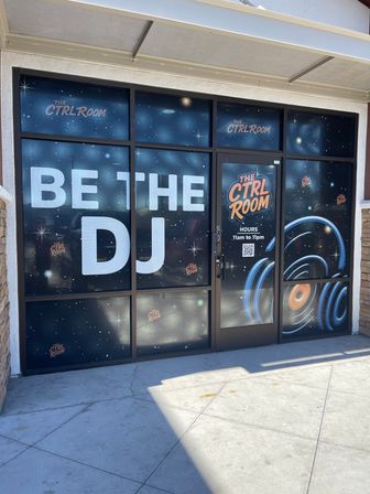 Strip-mall storefront with space-themed window graphics, large 'BE THE DJ' lettering and DJ vinyl/soundwave artwork on the glass door, sunlit sidewalk in front.