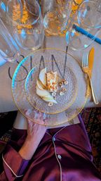 Top-down view of an elegant table setting: textured glass plate with gold rim holding a seared cheese canapé, sliced pear and hazelnuts, surrounded by stemless wine glasses and gold cutlery on a kraft-paper placemat.