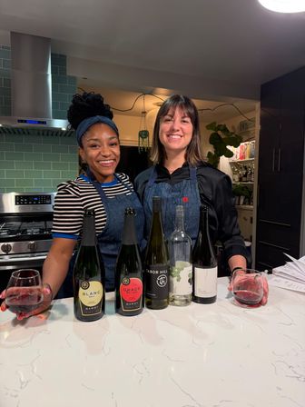 Two smiling women in aprons doing a home wine tasting at a marble kitchen island with several wine bottles on the counter, modern kitchen with teal subway tile and stainless range hood.