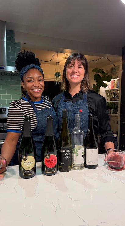 Two smiling women in aprons doing a home wine tasting at a marble kitchen island with several wine bottles on the counter, modern kitchen with teal subway tile and stainless range hood.