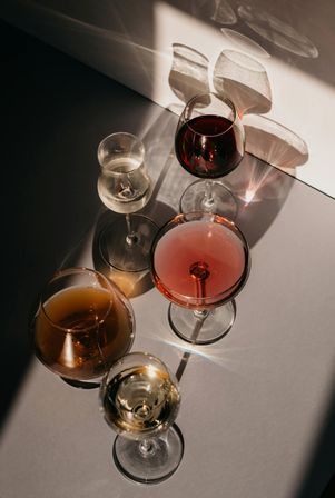 Top-down shot of five wine glasses—red, rosé, amber and two whites—on a sunlit tabletop, casting dramatic shadows and sparkling reflections in a stylish wine-tasting still life.