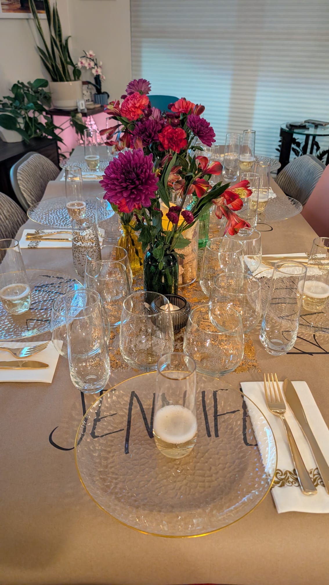 Vibrant home dining tablescape with magenta and red floral centerpieces in small vases, textured clear glass chargers and multiple wine glasses, gold flatware and candlelight on a kraft-paper runner.
