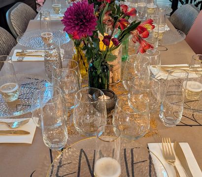 Vibrant home dining tablescape with magenta and red floral centerpieces in small vases, textured clear glass chargers and multiple wine glasses, gold flatware and candlelight on a kraft-paper runner.